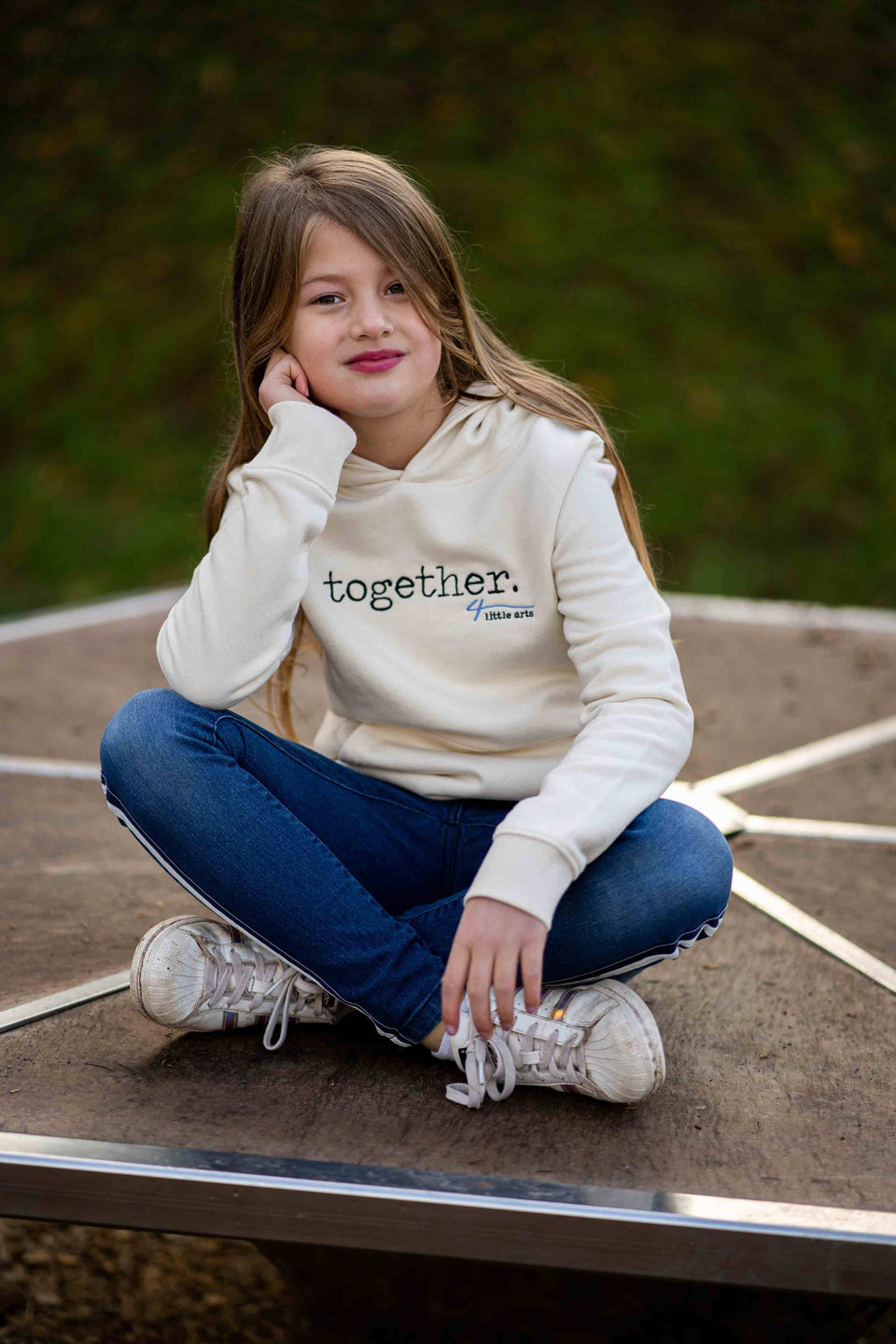 Ein Kindermodel trägt den cremefarbenen Kapuzenpullover mit gesticktem Schriftzug 'together' auf der Brust.