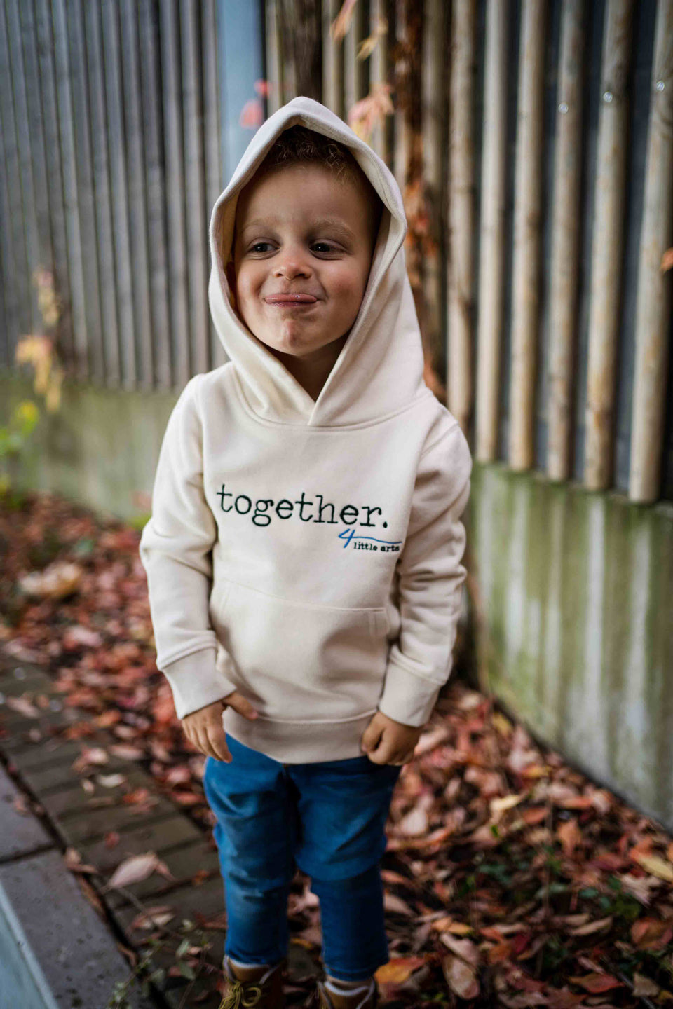Ein kleiner Junge streckt die Zunge raus und trägt den cremefarbenen Hoodie für Kinder mit dem gestickten Schriftzug 'together' auf der Brust.