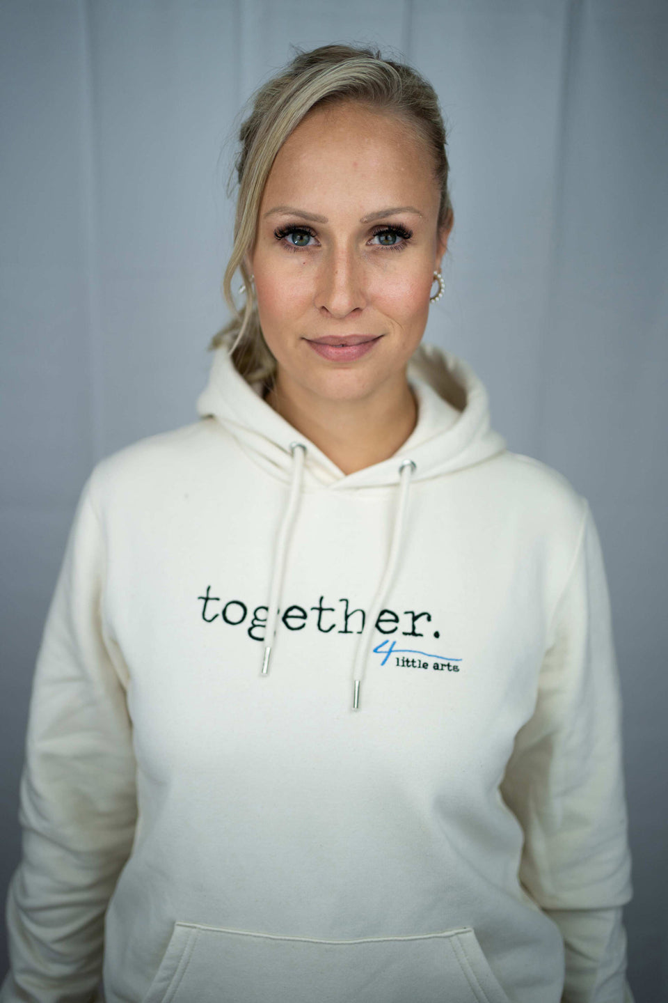 Ein weibliches Model trägt den cremefarbenen Unisex-Hoodie mit dem gestickten Schriftzug 'together' auf der Brust.