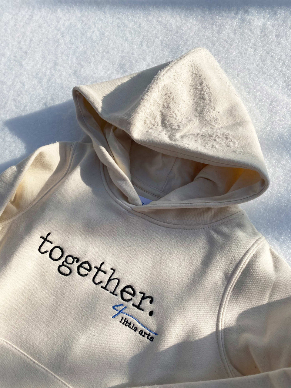Der cremefarbene Hoodie mit gesticktem 'together'-Schriftzug liegt im Schnee.