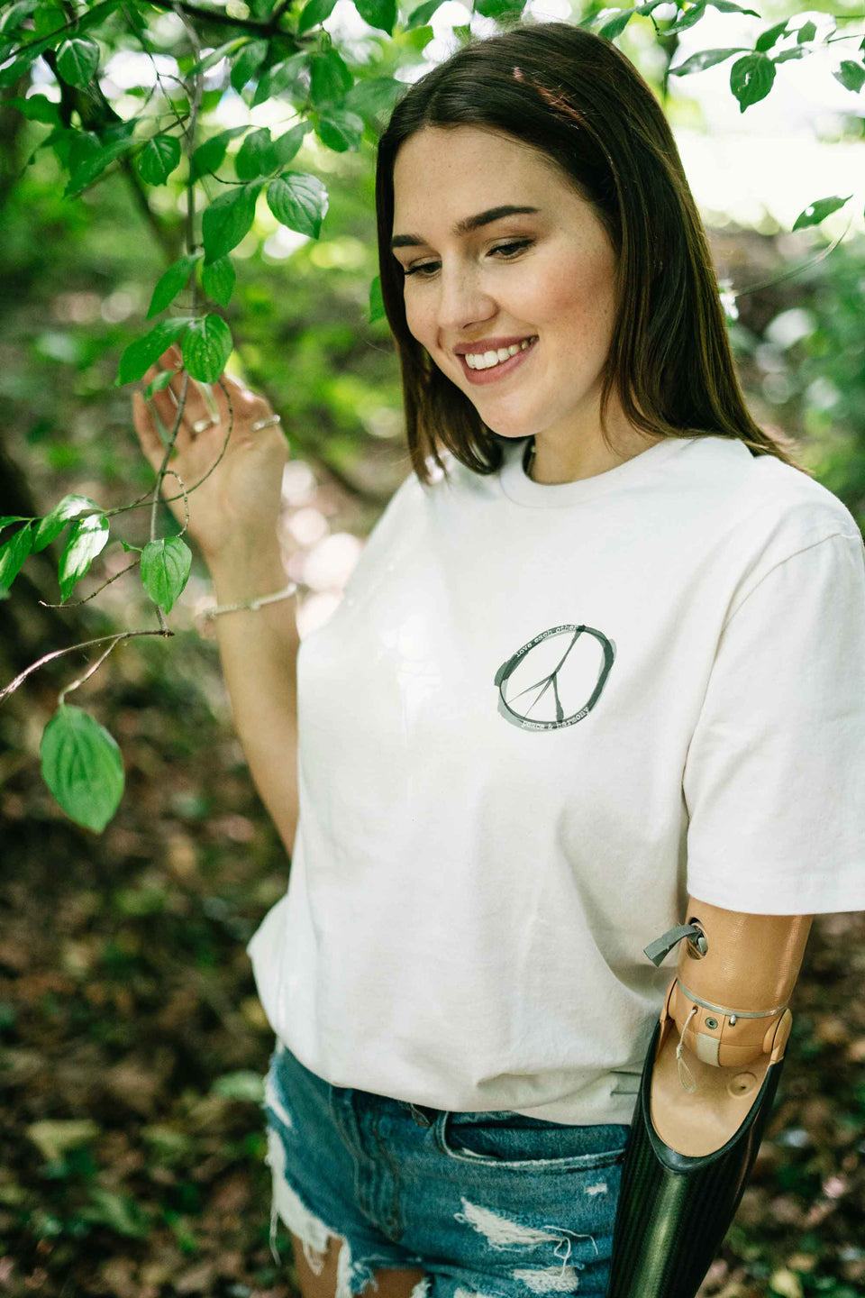 Ein Model trägt ein Unisex-T-Shirt aus recycelten Materialien mit dem Brustdruck 'Peace'. Das Shirt ist cremefarben, der Druck ist grau-schwarz.